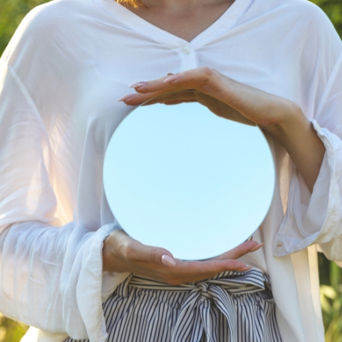 a woman in a white shirt holding a mirror infront of her chest reflecting the sky